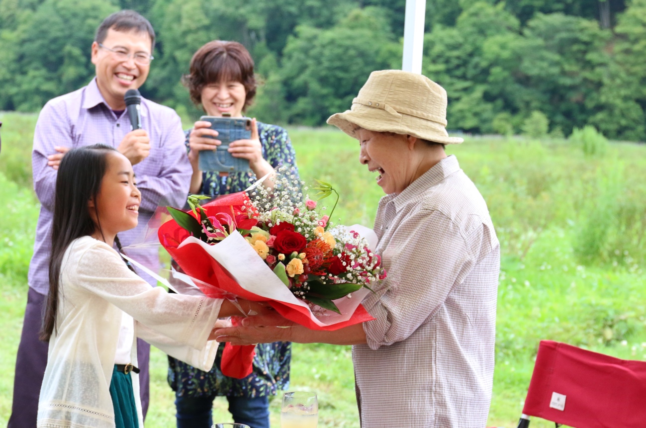 <p>お孫さんから美紀子さんへ花束の贈呈。山口さん夫婦が見つめます</p>